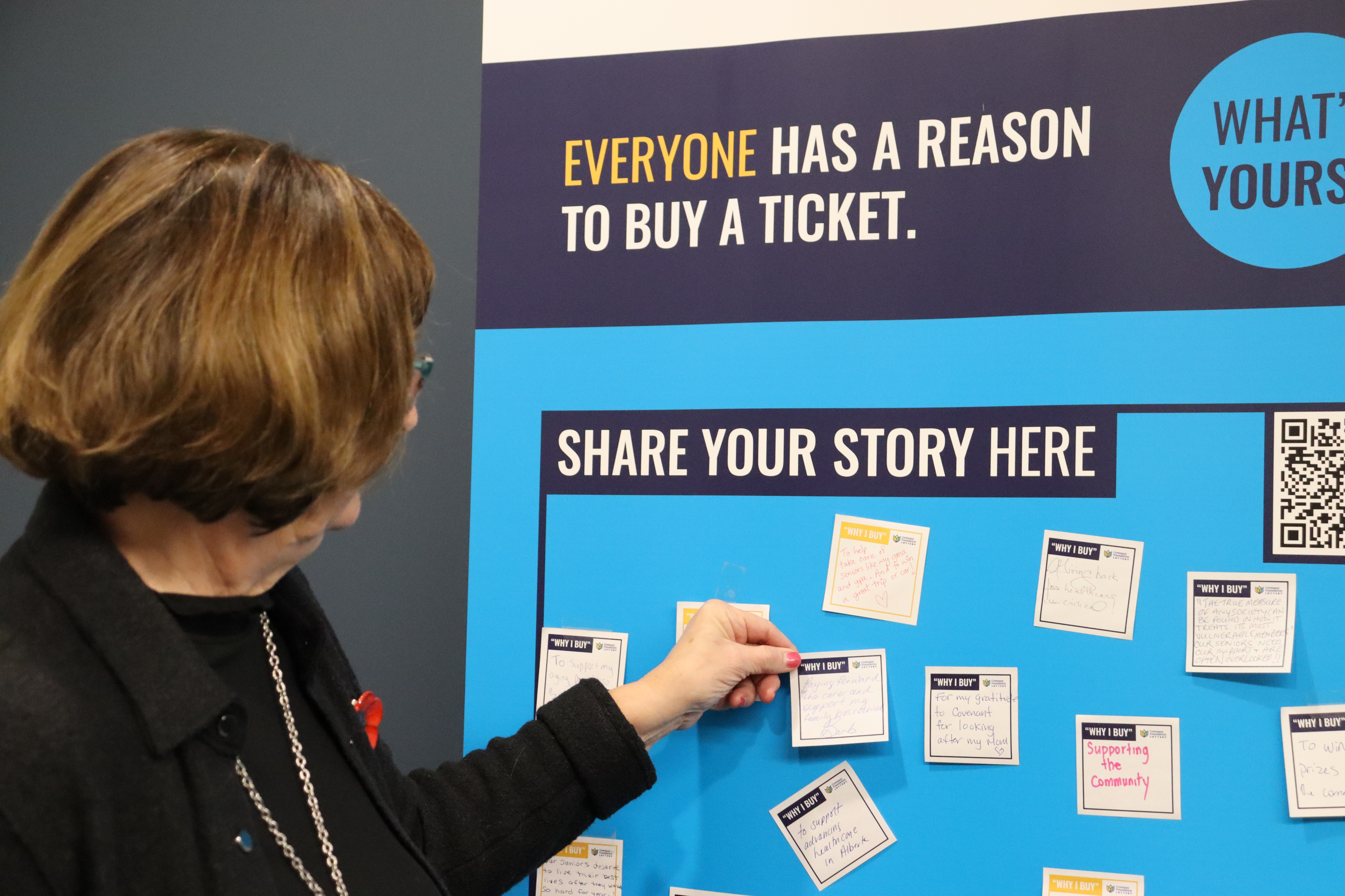 Barbara puts a post-it note on the Covenant Foundation Lottery banner stating why she buys tickets. 